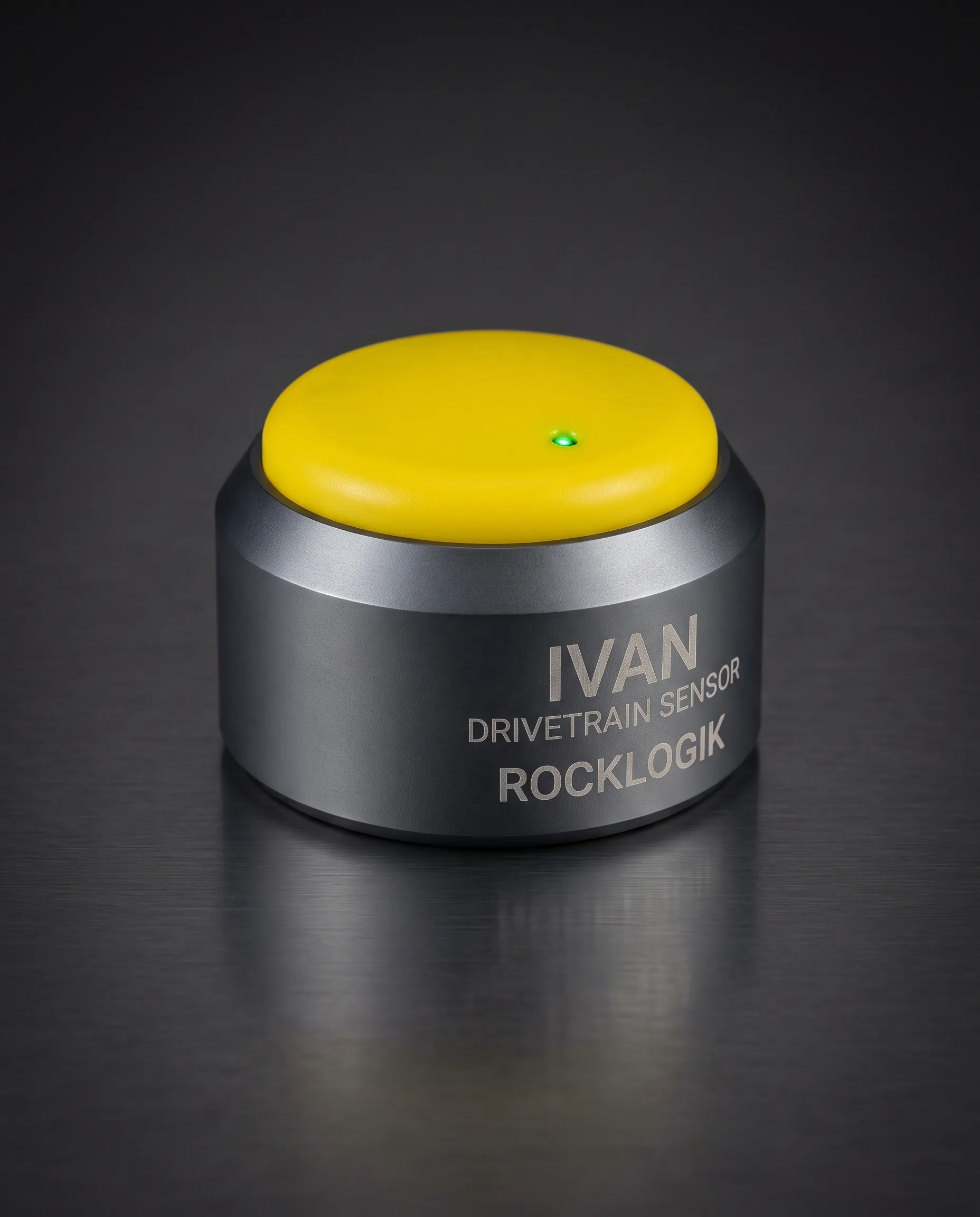 Studio photograph of the IVAN drivetrain sensor: a squat dark-grey aluminium puck with a domed yellow cap and a green status light, sitting flat on its base, etched IVAN, drivetrain sensor, RockLogik.